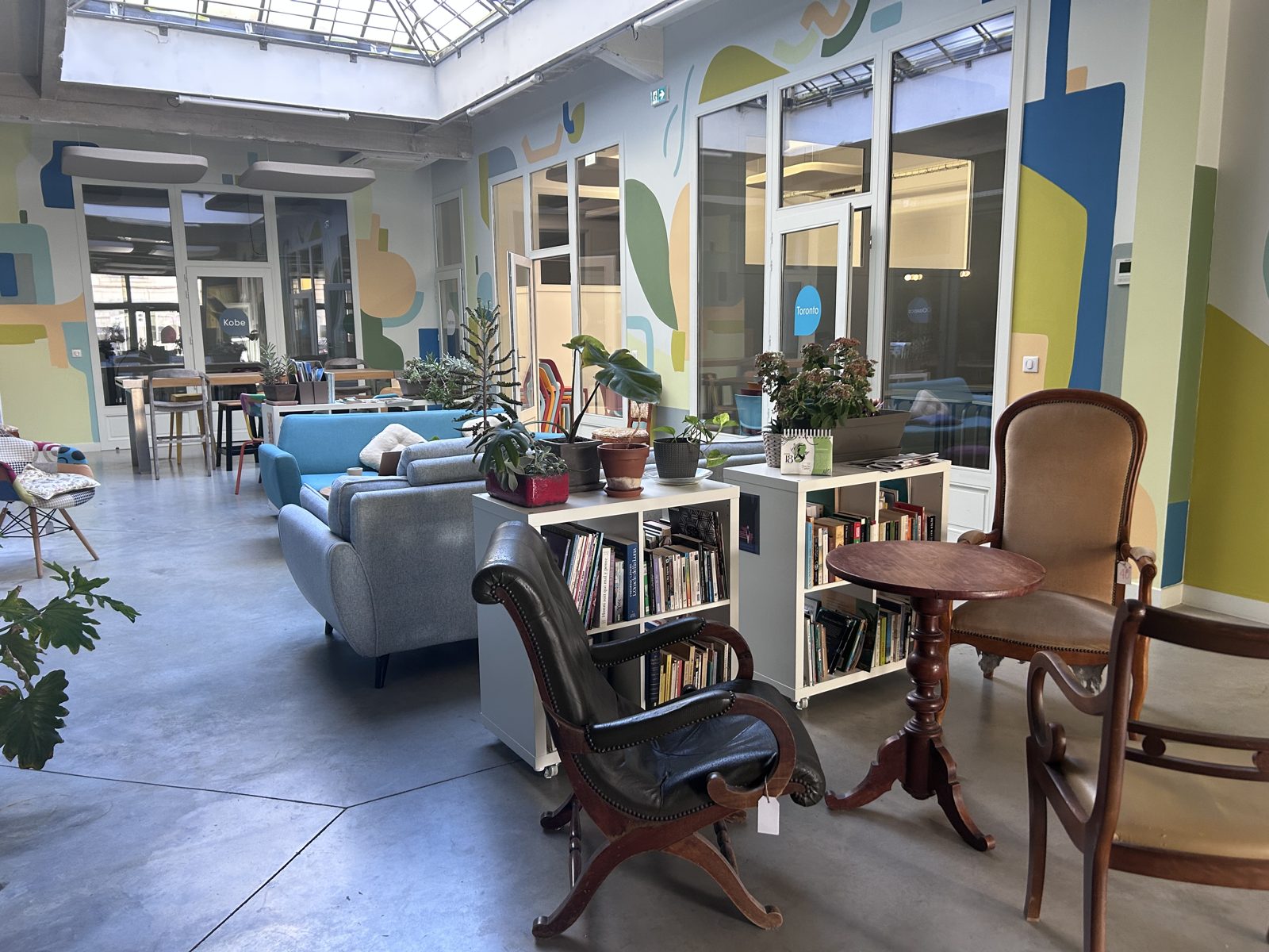 L’espace commun de Jakinola sous une grande verrière : canapés, fauteuils, bibliothèques, plantes et murs peints aux couleurs vives, avec les salles vitrées en fond.